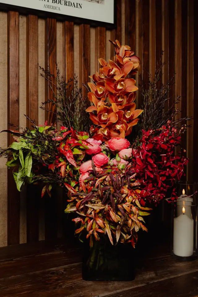 A vibrant, large floral arrangement is prominently displayed on a dark wooden table. It features a rich mix of orange orchids, pink ranunculus, and dark red flowers, complemented by various green and reddish foliage. Two lit candles in glass holders provide a warm, ambient glow to the right of the arrangement.