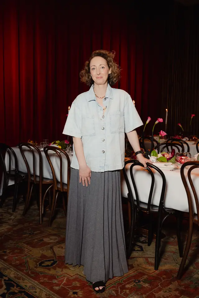 A woman with curly brown hair, wearing a light blue denim-style shirt and a grey pleated maxi skirt, stands smiling in front of a long dining table. She is positioned to the left of the frame, with her right hand resting on the back of a dark wooden chair. In the background, rich red curtains provide a dramatic backdrop, while the foreground features a patterned rug.