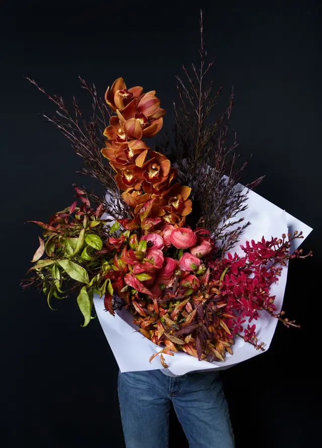 A large, vibrant floral bouquet is prominently displayed against a dark background. It features a striking arrangement of orange orchids, pink ranunculus, and a variety of green, red, and brown foliage, all wrapped in white paper. The bouquet is held by a person, with only their lower torso and jeans visible at the bottom of the frame.