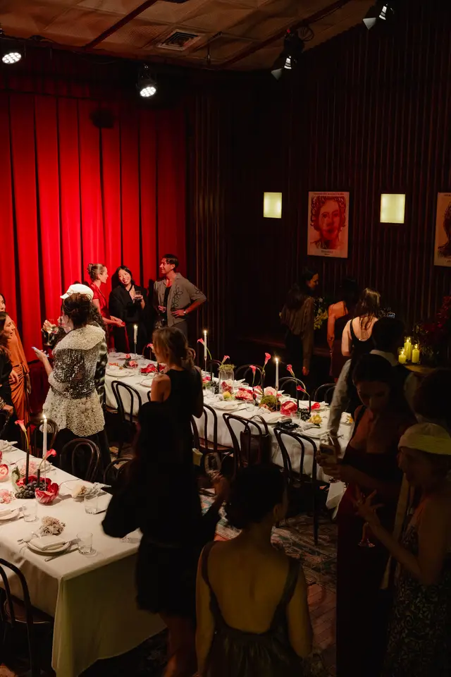 A vibrant indoor event scene captures guests mingling around elegantly set dinner tables. The room features dramatic red curtains, dark wood paneling, and warm, intimate lighting from spotlights and numerous candles. Tables are adorned with white tablecloths, elaborate floral arrangements, and fresh fruit, creating a sophisticated and festive atmosphere.