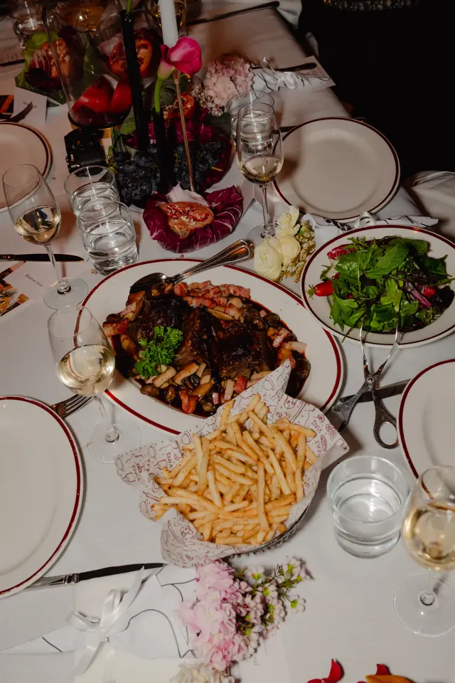 A top-down view of an elegantly set dining table, showcasing a lavish meal and decorative elements. The table is adorned with a white linen tablecloth, multiple place settings, a main course, side dishes, wine, water, and elaborate floral and fruit centerpieces, suggesting a formal event or upscale restaurant setting.