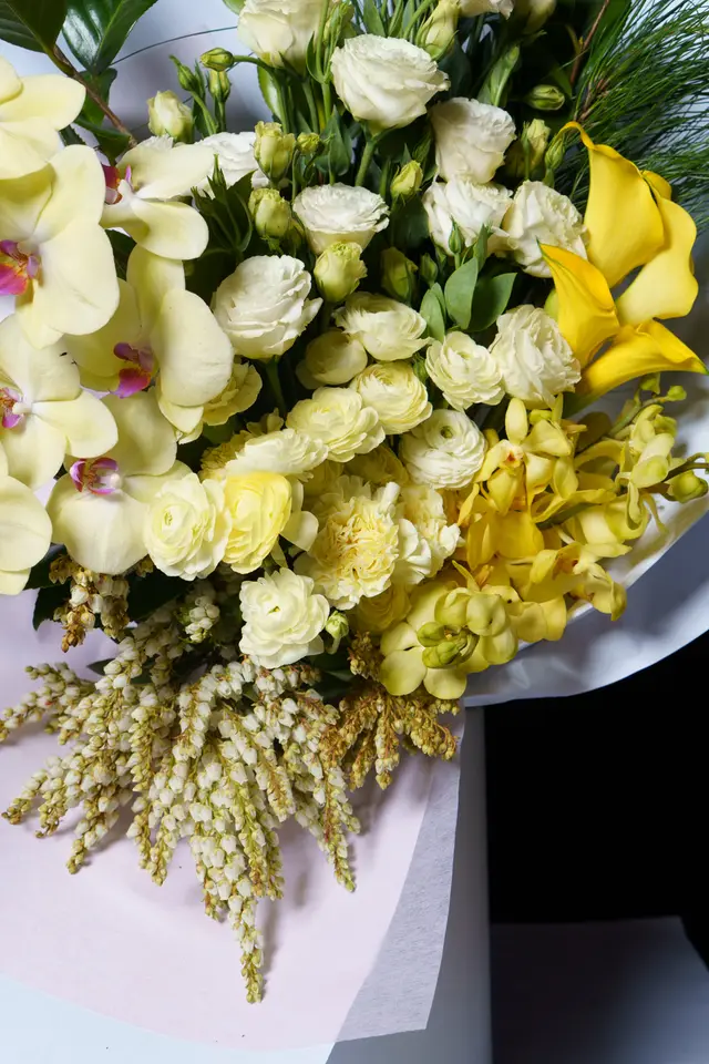 A vibrant, close-up studio shot of a large floral bouquet featuring a variety of yellow and white flowers. The arrangement is artfully composed with different textures and shades, wrapped in light-colored paper. The lighting highlights the fresh details of each bloom.