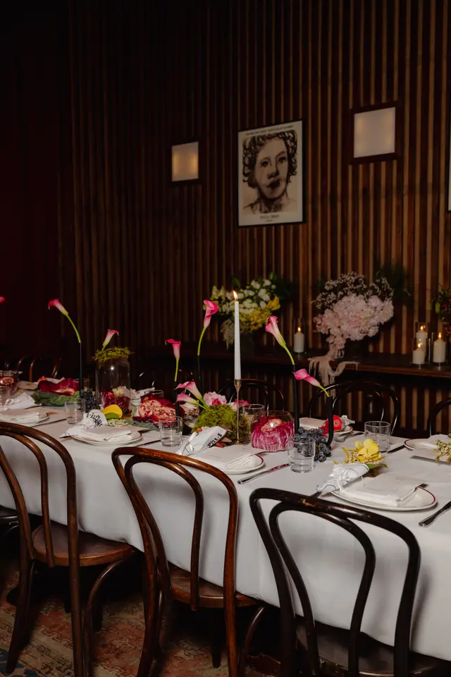 A long dining table is elegantly set for an event, featuring multiple place settings with white tablecloths, plates, and glassware. The table is adorned with various floral arrangements, lit candles, and decorative elements like pomegranates, grapes, and lemons. The background shows a dark wooden slatted wall with framed artwork and wall sconces, creating a warm and intimate atmosphere.