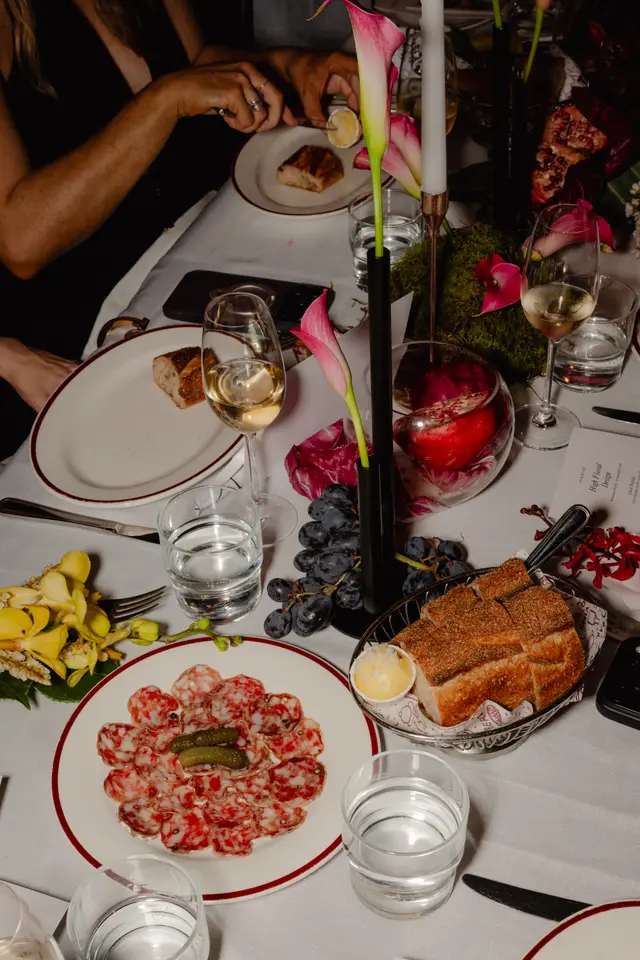 A high-angle, close-up shot captures an elegantly set dinner table at an event. The table features various dishes including sliced salami with gherkins, a bread basket with butter, and a bunch of dark grapes. Floral arrangements with pink calla lilies and yellow orchids, a lit candle, and a decorative pomegranate enhance the sophisticated ambiance, with hands visible interacting with the setting.