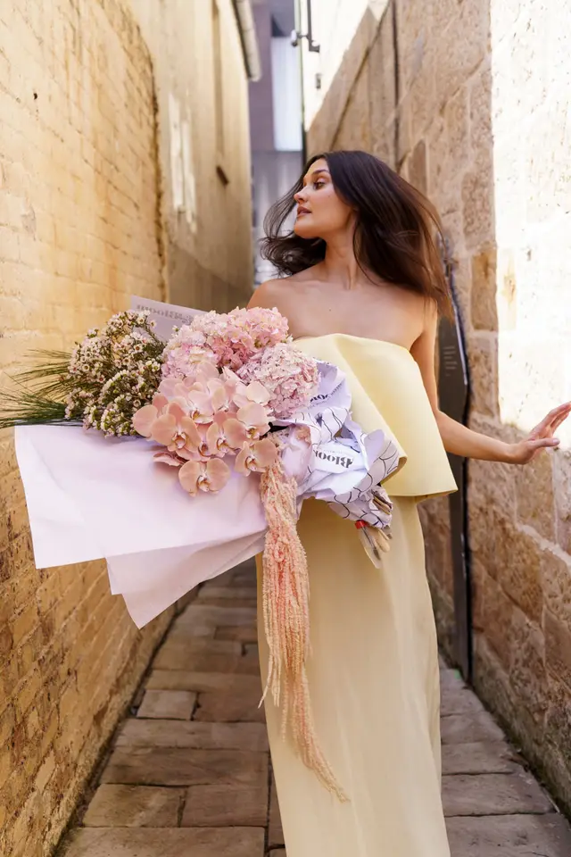 A full-length shot of a woman in a light yellow strapless dress, holding a large, pastel-colored floral bouquet. She is walking through a narrow outdoor alleyway with old brick walls, looking to her right with her dark hair flowing. The bouquet features various pink and peach flowers wrapped in white and patterned paper.