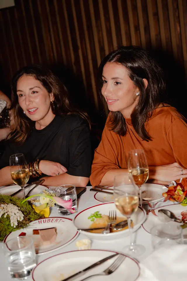 Two women are seated at a dinner table, engaged in conversation or listening intently. The woman on the left wears a black top and gold bangles, while the woman on the right wears an orange-brown top and a ring. The table is set with white tablecloths, plates, cutlery, wine glasses, and water glasses, along with some floral and mossy decorations.