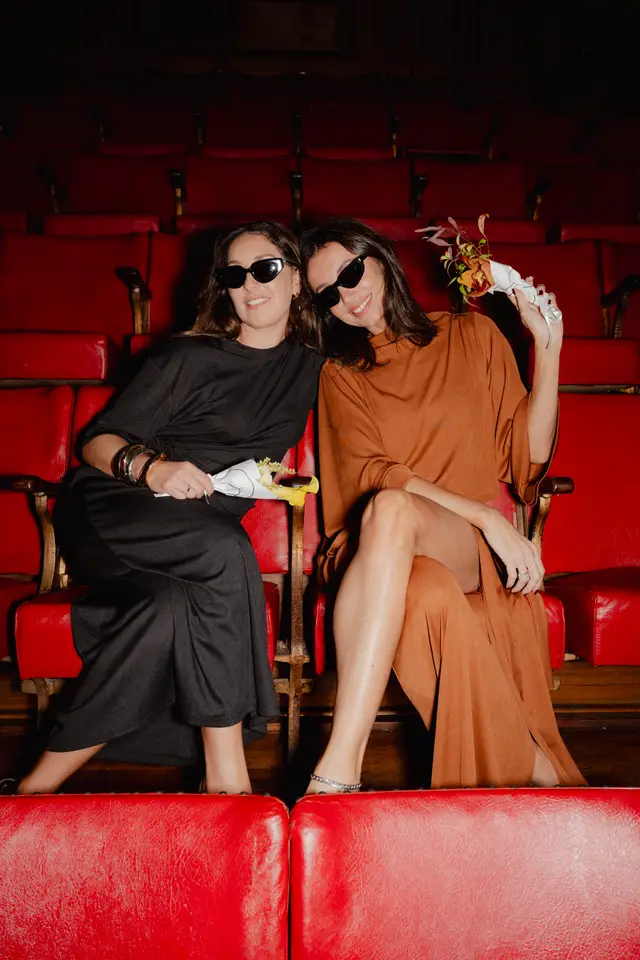 Two women, both wearing sunglasses and long dresses, are seated side-by-side in a row of red theater seats. They are smiling and each holds a small, artfully arranged floral bouquet wrapped in white paper. The background shows more rows of empty red seats, suggesting a theater or auditorium setting.