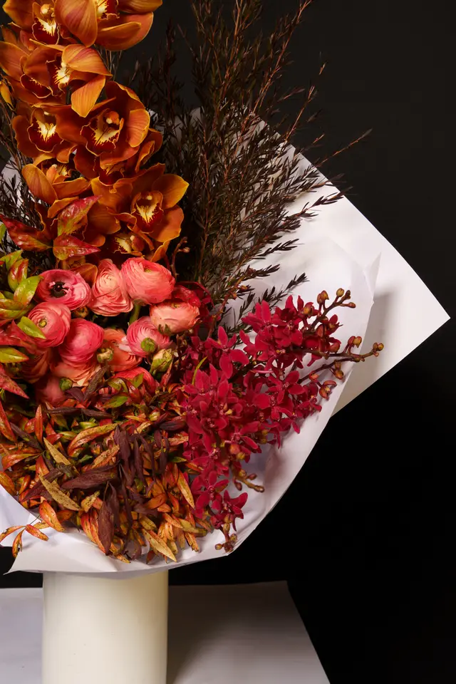 A vibrant, professionally arranged floral bouquet featuring a mix of orange orchids, coral ranunculus, and deep red orchids, complemented by various autumnal foliage. The bouquet is wrapped in white paper and presented on a white cylindrical stand against a stark black background, emphasizing its rich colors and textures. The composition is a close-up, vertical shot highlighting the intricate details of the flowers and leaves.