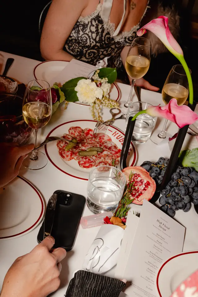 A close-up, overhead view of an elegantly set dining table at an event. The table features white tablecloths, red-rimmed plates, wine and water glasses, and various food items including charcuterie, grapes, and pomegranate. Decorative floral arrangements with pink calla lilies and white flowers are interspersed among the place settings, with hands of multiple diners visible interacting with the table.