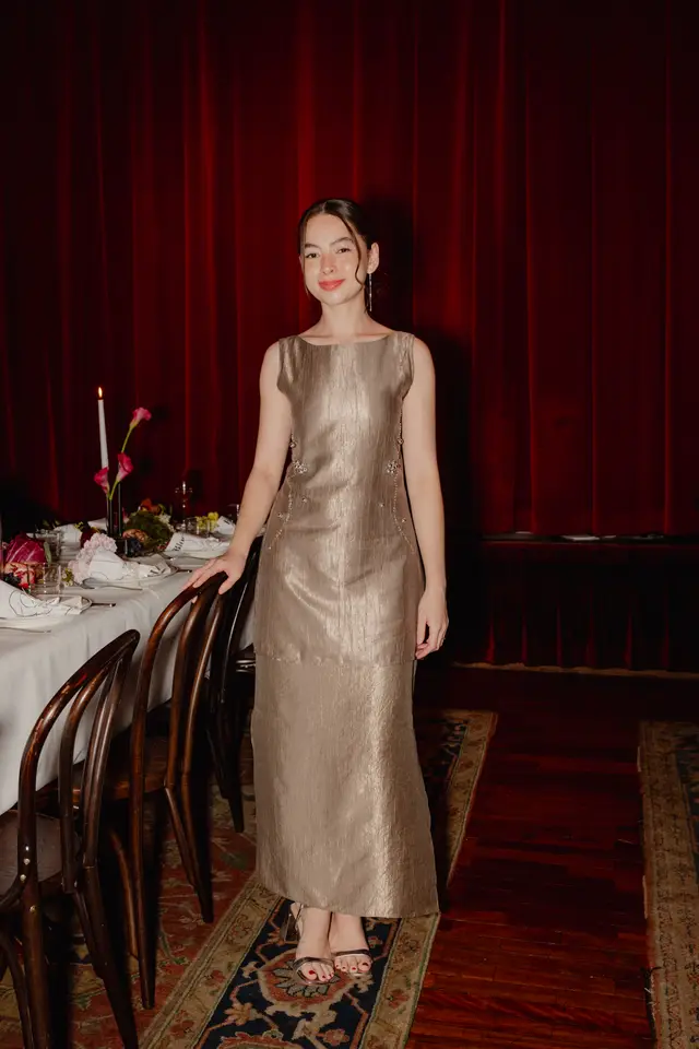 A young woman in a shimmering gold dress stands smiling next to a beautifully set dinner table. The background features rich red velvet curtains, suggesting a formal event or celebration. The table is adorned with a white tablecloth, dark wooden chairs, a lit candle, and decorative floral and fruit arrangements.