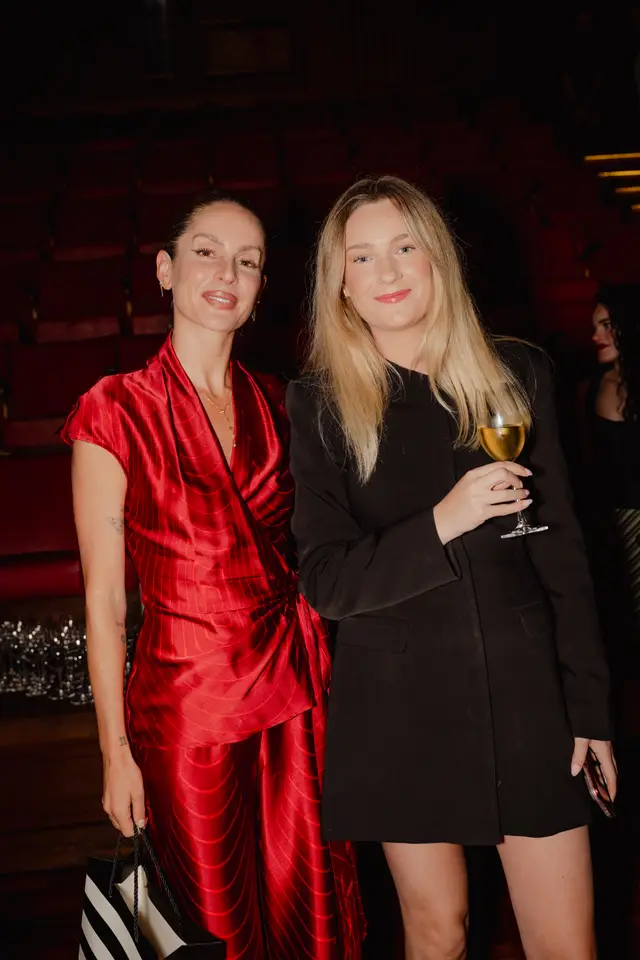 Two women pose and smile directly at the camera in a dimly lit indoor setting. The woman on the left wears a red patterned outfit and holds a black and white striped shopping bag, while the woman on the right wears a black blazer dress and holds a glass of white wine. Rows of red theater-style seating are visible in the blurred background.