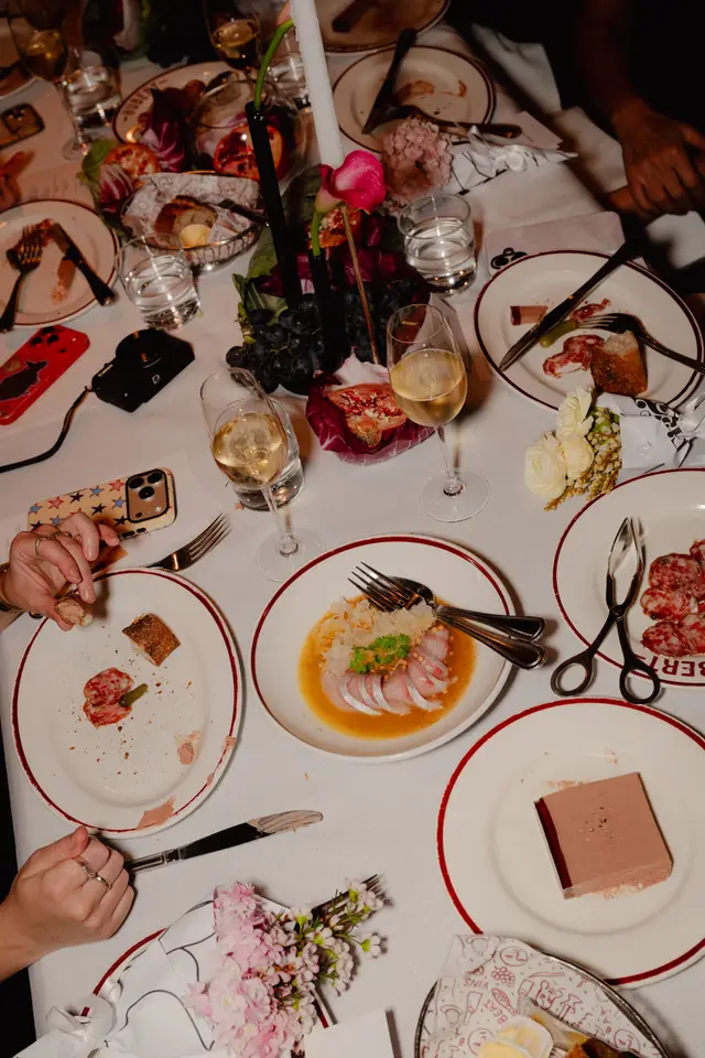 An overhead shot of a lively dinner table at an event, featuring various dishes, wine glasses, and decorative floral arrangements. Several hands are visible, indicating multiple guests enjoying the meal and conversation. The table is set with white linen and plates with red rims, creating an elegant and festive atmosphere.