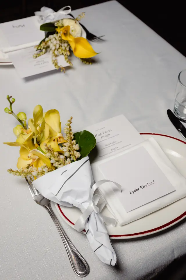 A close-up, overhead shot of an elegant table setting featuring a white tablecloth, a white plate with a red rim, and a silver fork. A small bouquet wrapped in white paper with yellow orchids and white bell flowers is placed on the plate. A menu card for 'High Floral Design' and a place card for 'Lydia Kirkland' are also visible, along with a clear glass and a second, smaller floral arrangement in the background.