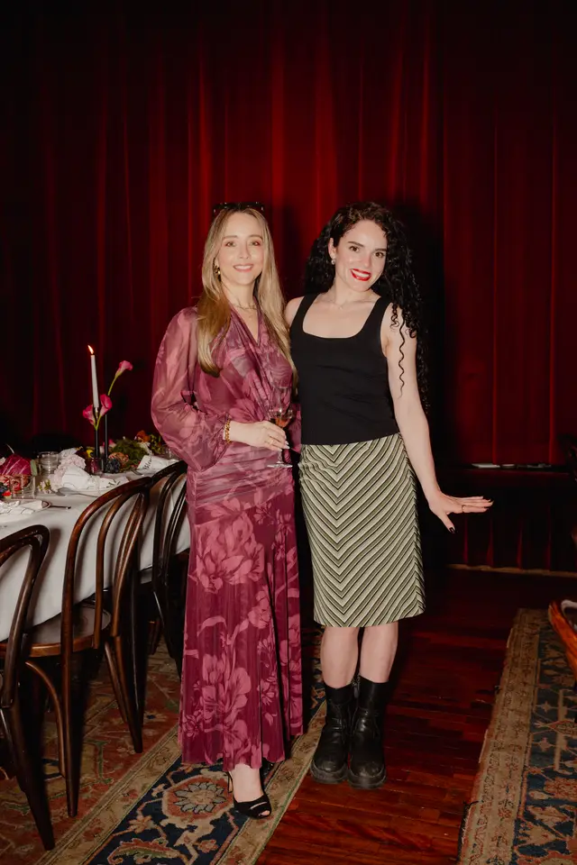 Two women are posing for a full-body portrait at an indoor event. They stand in front of a long dining table set with a white tablecloth, flowers, and a lit candle, with deep red velvet curtains forming the background. The woman on the left wears a long, sheer burgundy floral dress and holds a glass of rosé wine, while the woman on the right wears a black tank top, a green and white striped skirt, and black boots.