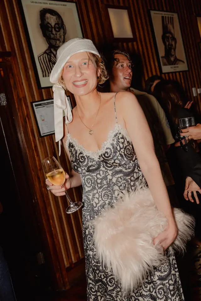 A smiling woman in a white bucket hat and a black and white patterned slip dress holds a glass of white wine and a light pink fluffy clutch. She is positioned centrally, with a warm, dimly lit background featuring wood-paneled walls and framed black and white portraits. Other guests are visible out of focus behind her, suggesting a social event.