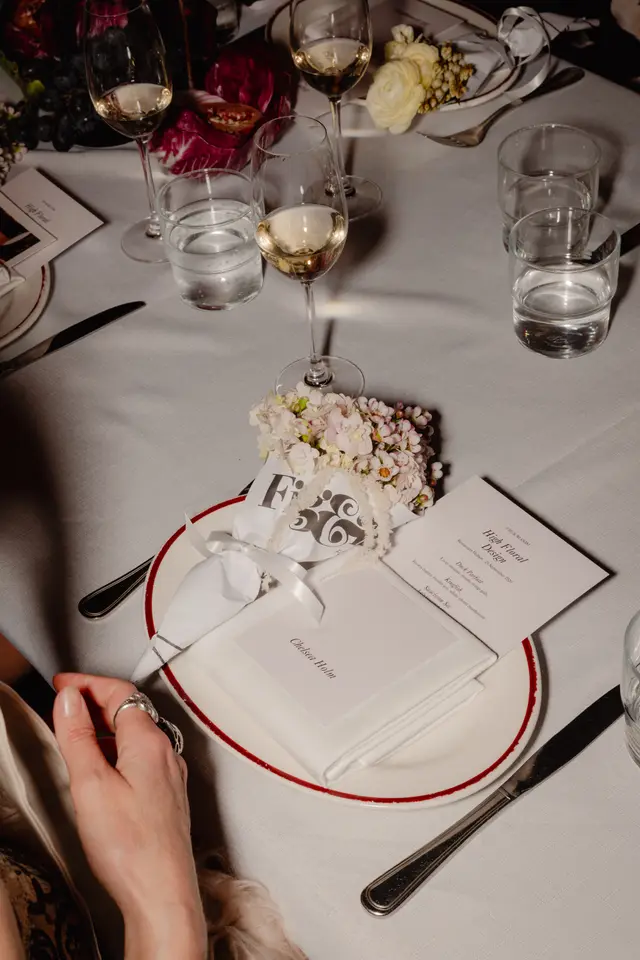 A close-up, overhead shot of an elegant table setting at an event, featuring a white plate with a red rim, a personalized place card, a menu for a 'High Floral Design' event, and a delicate floral arrangement. Wine and water glasses, along with cutlery, are also visible on the crisp white tablecloth, with a person's hand resting near the plate.