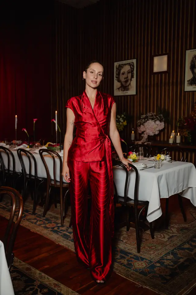 A woman in a vibrant red patterned wrap top and matching wide-leg pants stands beside a long dining table set for an event. She is looking directly at the camera, with one hand resting on a dark wooden chair. The background features warm lighting, floral arrangements, candles, and framed artwork on a wood-paneled wall.