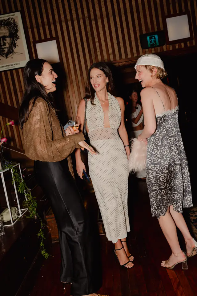 Three women are captured mid-conversation and laughter at an indoor event, illuminated by warm, dim lighting. They are dressed in fashionable attire, holding drinks, and appear to be enjoying a lively social gathering. The background features a distinctive striped wall and subtle floral decor.
