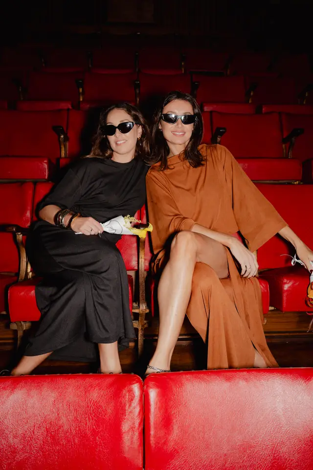 Two women wearing sunglasses and stylish dresses are seated side-by-side in a dimly lit theater with rows of vibrant red seats. They are looking directly at the camera with subtle smiles. The woman on the left holds a small bouquet of white and yellow flowers, while the woman on the right holds a white-wrapped item.