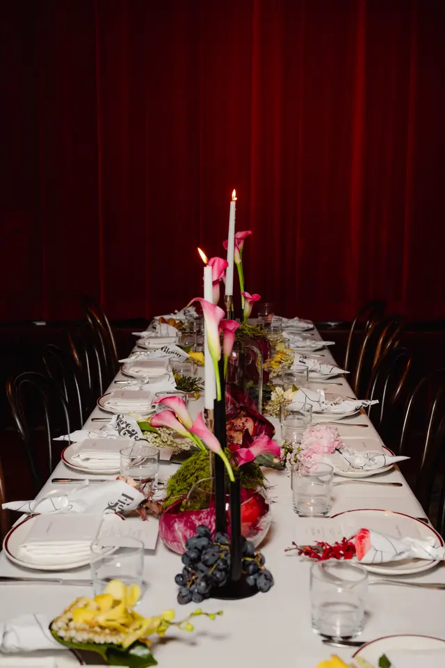A long dining table is elegantly set for an event, featuring multiple place settings, elaborate floral arrangements, and lit candles. The table is adorned with pink calla lilies, yellow orchids, and various fruits, set against a backdrop of deep red curtains. The scene conveys a sophisticated and inviting atmosphere.