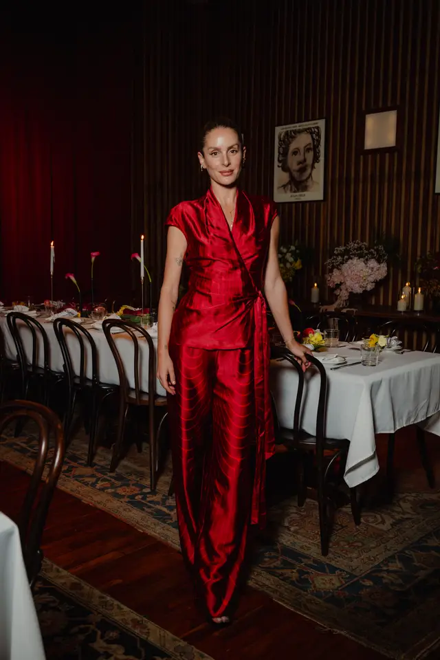 A woman in a vibrant red patterned silk wrap top and matching wide-leg pants stands in a dimly lit event space. She is positioned next to a long dining table set with white tablecloths, lit candles, and floral arrangements, with dark wood paneling and red curtains in the background.