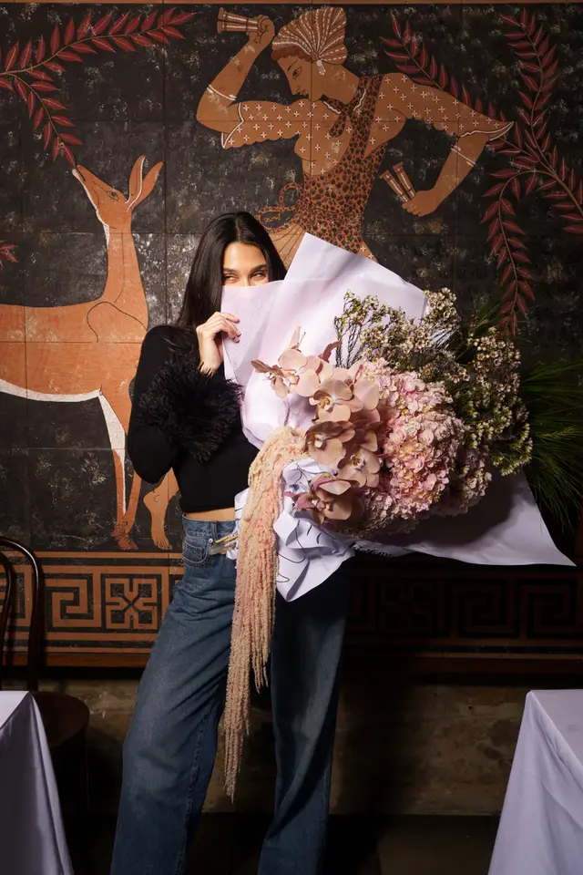 A woman with long dark hair holds a large, vibrant bouquet of flowers, partially obscuring her face with the light purple wrapping paper, revealing only her eyes. She wears a black top with feathered cuffs and blue jeans, standing against a dark wall adorned with ancient-style murals featuring a deer and a human figure. The setting includes a dark wooden chair and a white-draped table, suggesting an indoor location.
