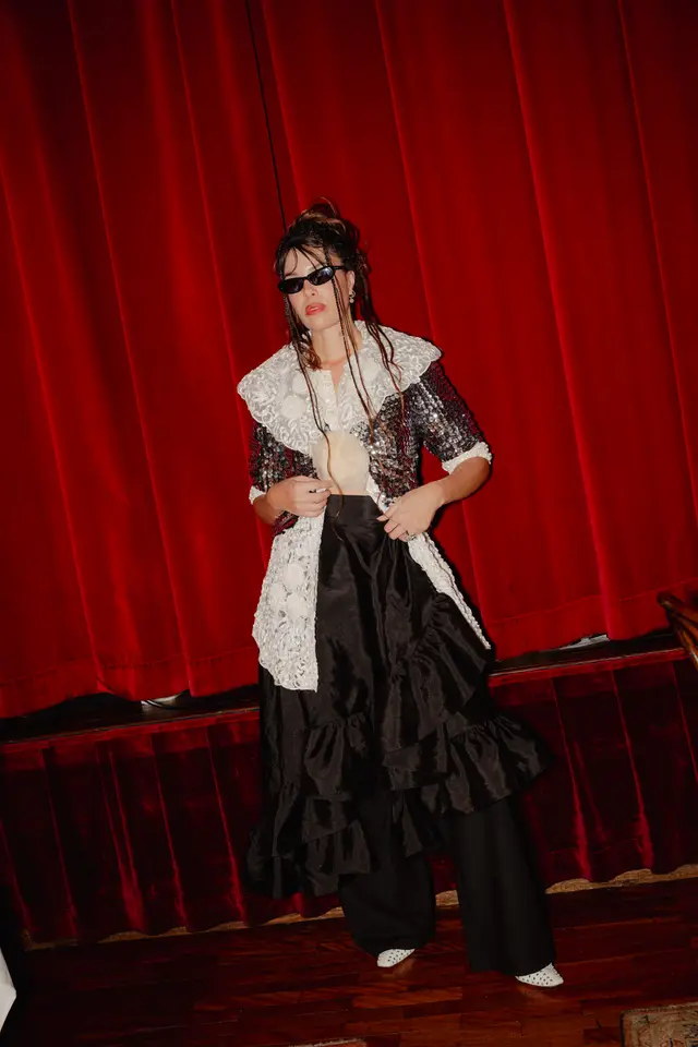 A woman in a striking, full-length outfit stands against a deep red velvet curtain backdrop. She wears black sunglasses, a sequined jacket with a large white lace collar, a ruffled black skirt over black trousers, and white studded shoes. Her hands are positioned near her waist, and her hair is styled with braids.