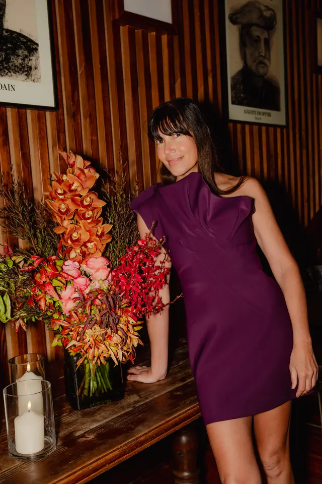 A woman in a vibrant purple one-shoulder dress smiles at the camera, leaning on a dark wooden table. Beside her is a large, colorful floral arrangement featuring orange orchids, pink ranunculus, and various red and green foliage. The background consists of a warm-toned slatted wooden wall adorned with framed black and white artwork, and two lit candles in glass holders are visible on the table.