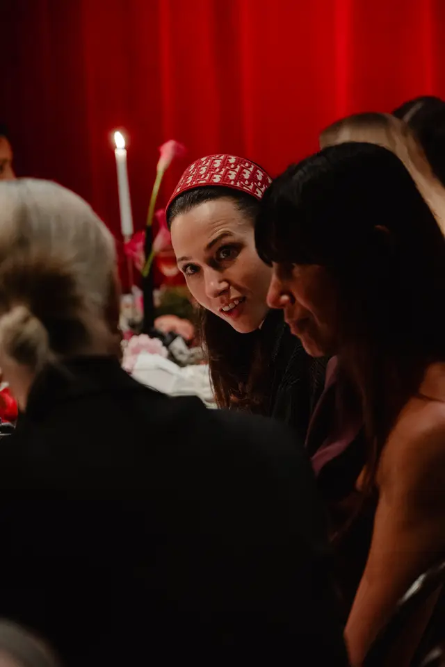 A close-up, intimate shot of a woman wearing a red and white patterned headband, looking directly at the viewer with a slight smile. She is seated at a dimly lit table with a lit candle and pink flowers, surrounded by other guests. The background features soft red drapes, suggesting an elegant evening event.