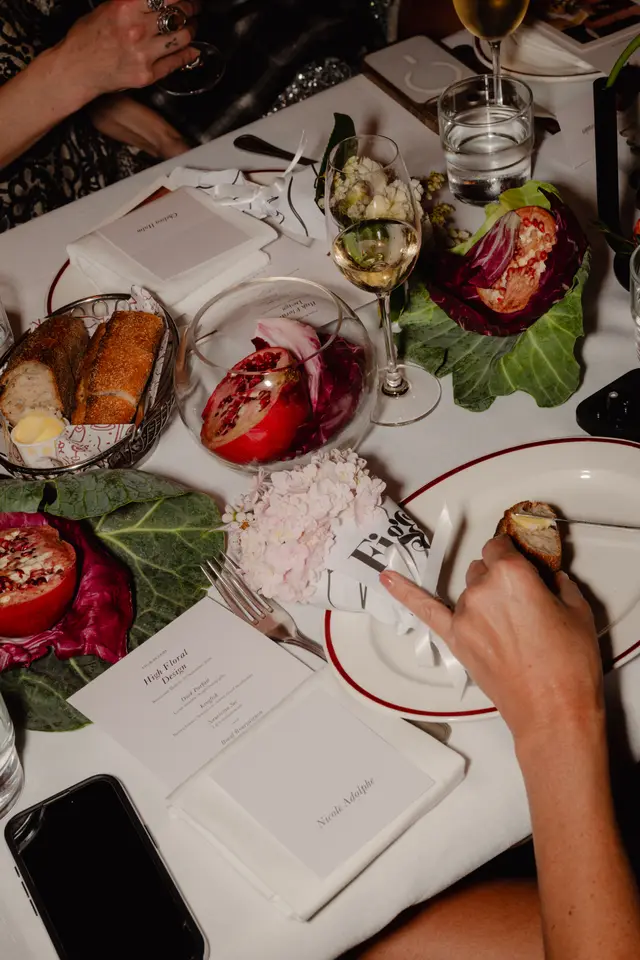 An overhead shot captures a beautifully set dinner table, featuring various dishes, drinks, and decorative elements. Hands are visible interacting with the food and table items, suggesting an active dining experience. The scene includes bread, butter, wine, water, pomegranates, and floral arrangements, all arranged on a white tablecloth.