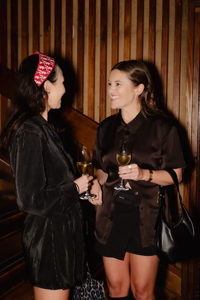Two women are captured mid-conversation, smiling and holding glasses of white wine. They are dressed in stylish dark attire, standing against a warm, vertically slatted wooden wall. The image conveys a relaxed and social atmosphere at an event.