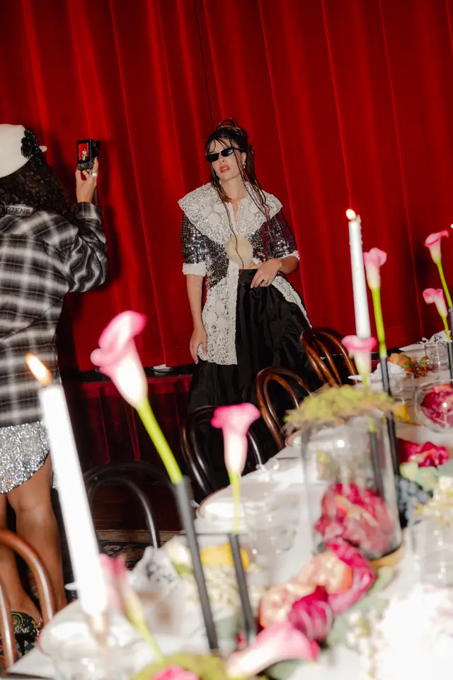 A vertical shot captures a person in a sequined and lace outfit, wearing sunglasses, posing in front of a deep red curtain backdrop. Another person is visible from behind, holding a camera to photograph the subject. In the foreground, a table is set with white tablecloths, dark wooden chairs, lit candles, and arrangements of pink calla lilies and other decorative elements.