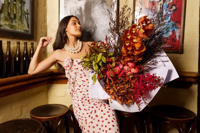 A woman with dark hair and a pearl necklace, wearing a white and red polka dot dress, leans against a wooden shelf. She holds a large, vibrant bouquet of orchids, ranunculus, and other foliage wrapped in white paper. The setting appears to be an indoor space with a cream-colored brick wall, framed artwork, and dark bottles on a shelf.