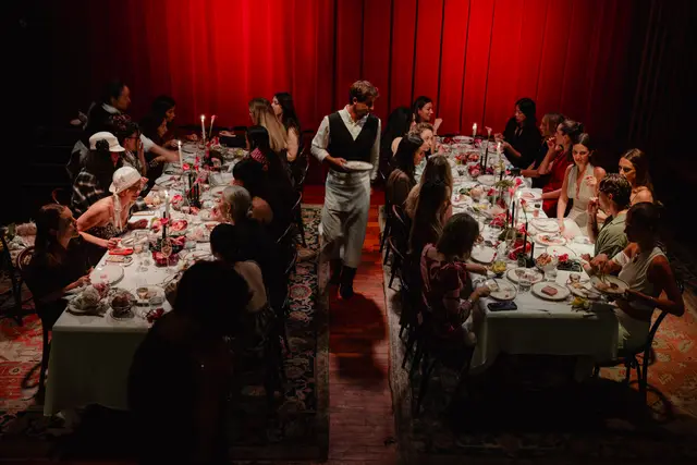 An overhead shot captures a long, candlelit dinner table filled with guests and elaborate floral and fruit arrangements, set against a dramatic red curtain backdrop. A server walks down the aisle between two rows of diners, carrying a plate. The scene is dimly lit, creating an intimate and sophisticated atmosphere for a social gathering.