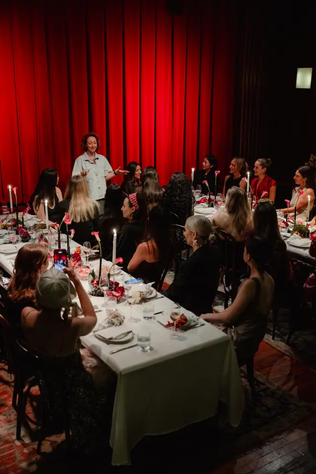 A woman in a light denim shirt speaks to a group of women seated around a long, white-clothed table. The table is adorned with candles, floral arrangements featuring pink calla lilies, and various place settings. A vibrant red curtain serves as the backdrop, creating an intimate and elegant atmosphere.