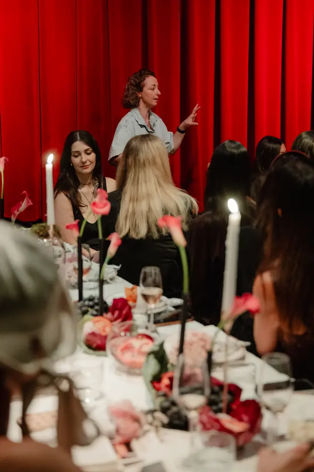 A speaker with curly hair and a light blue shirt addresses an audience seated at a long, elegantly decorated table. The background features rich red curtains, and the table is adorned with lit candles, pink and red flowers, grapes, and wine glasses, creating a warm and inviting atmosphere. The focus is on the interaction between the speaker and the attentive audience, with elements of a sophisticated event.