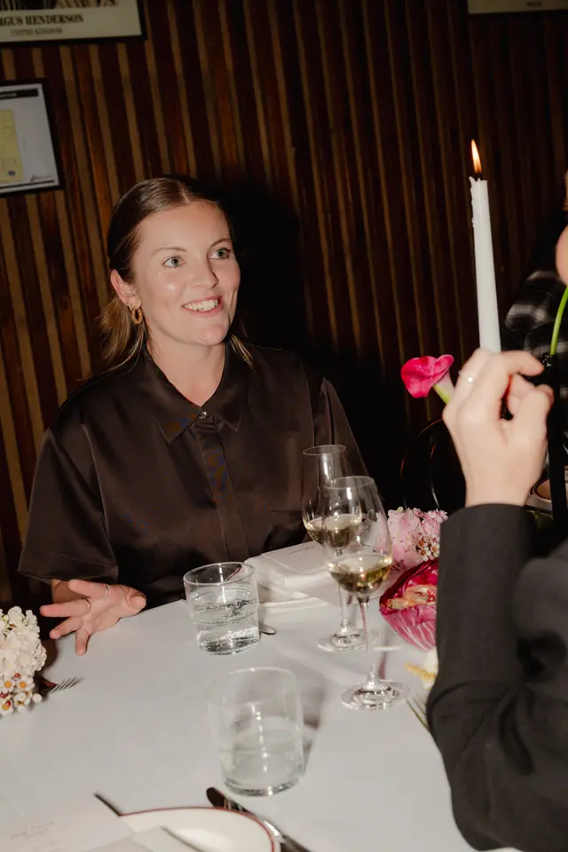 A smiling woman with light brown hair, wearing a dark brown collared shirt, is seated at a dinner table. The table is set with wine glasses, water glasses, white napkins, and small floral arrangements. A lit candle and a wooden slatted wall are visible in the background, suggesting an indoor event setting.