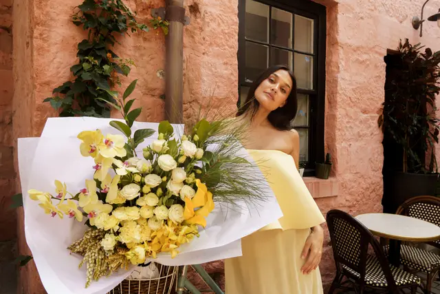 A woman in a strapless yellow dress stands next to a large, vibrant bouquet of yellow and white flowers, wrapped in white paper and resting in a bicycle basket. The scene is set outdoors against a textured reddish-pink wall with a dark-framed window and an outdoor cafe area visible in the background. The woman looks towards the camera with a gentle expression.