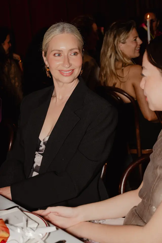 A blonde woman, wearing a black blazer over a black and white floral top, smiles while seated at a table. She is looking towards another person, partially visible in the foreground. The background is dimly lit with blurred figures, suggesting an evening event.