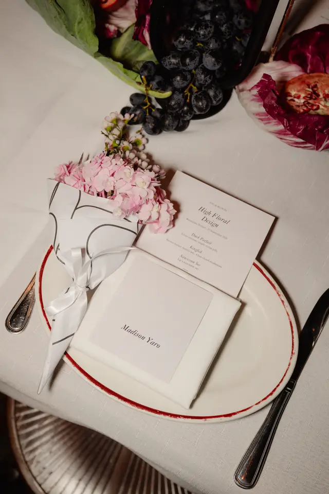 A close-up, overhead shot of an elegant table setting for an event. It features a white plate with a red rim, a small bouquet of pink flowers wrapped in patterned paper, a menu card for 'High Floral Design' by 'Fig & Bloom,' and a name card for 'Madison Yaro.' Decorative elements like dark grapes, green leaves, and radicchio are also visible on the white linen tablecloth.