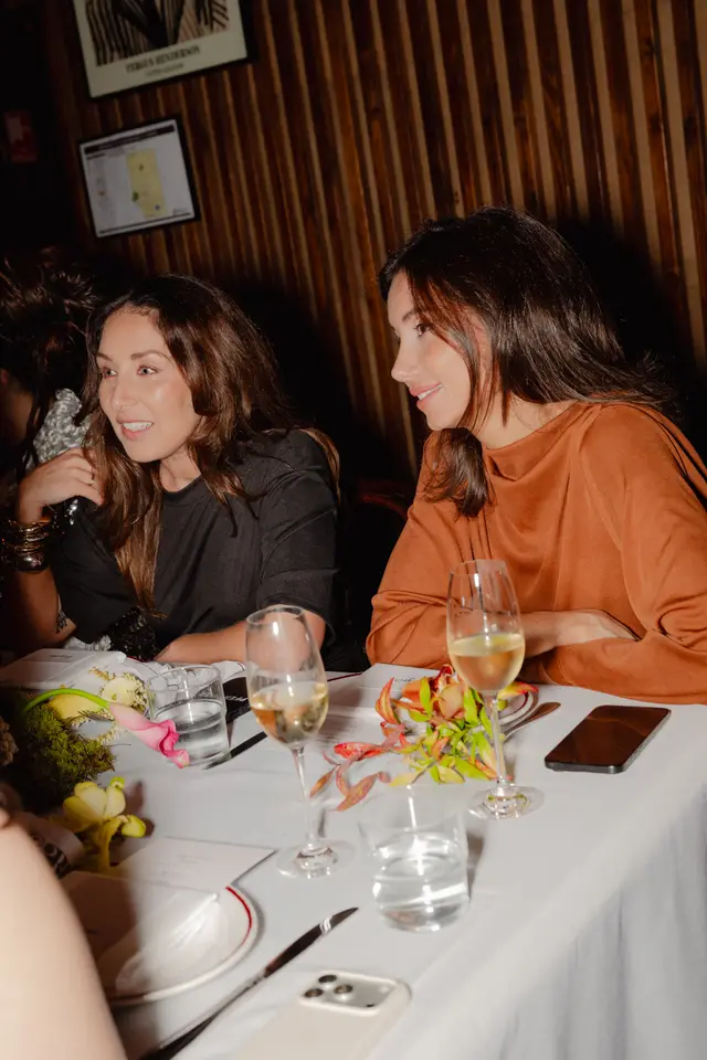 Two women are seated at a table, engaged in conversation at what appears to be an evening event. The table is set with drinks, cutlery, and elaborate floral arrangements featuring various flowers and foliage. The background shows a dark, warm-toned wooden wall with framed artwork, suggesting an upscale restaurant or event venue.