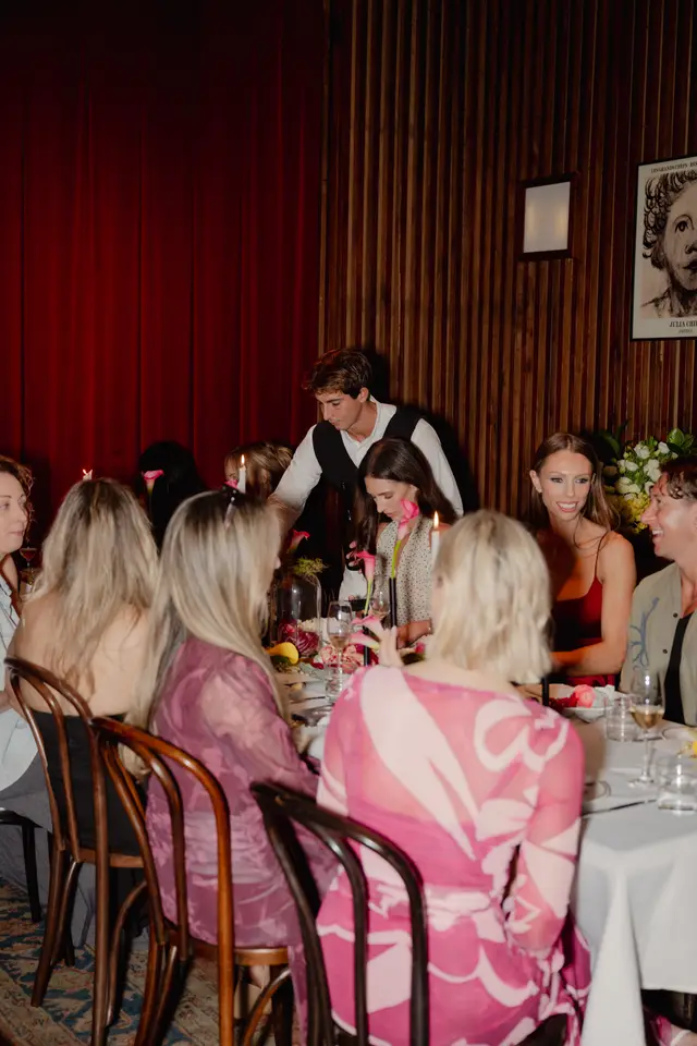 The image captures a lively dinner event with multiple guests seated around a table adorned with floral arrangements and fruit. A server attends to the guests, while warm lighting from candles and ambient fixtures illuminates the scene against a backdrop of red curtains and wood paneling. The atmosphere appears social and elegant, with people engaged in conversation.