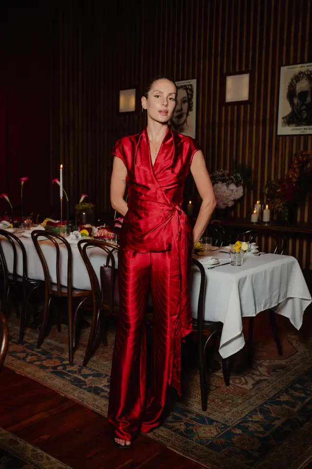 A full-length portrait of a woman in a vibrant red patterned wrap top and wide-leg pants, standing confidently in an elegantly decorated, dimly lit dining room. The background features a long table set for an event with white tablecloths, candles, and various floral arrangements, against a dark wood-paneled wall adorned with framed artwork.