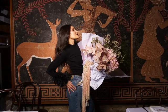 A woman with long dark hair, wearing a black ribbed top with feather cuffs and blue jeans, holds a large, elaborate bouquet of pink and peach flowers. She stands in profile against a dark wall mural depicting stylized ancient figures and animals. Dark wooden chairs and a partially visible white tablecloth suggest a dining or studio setting.