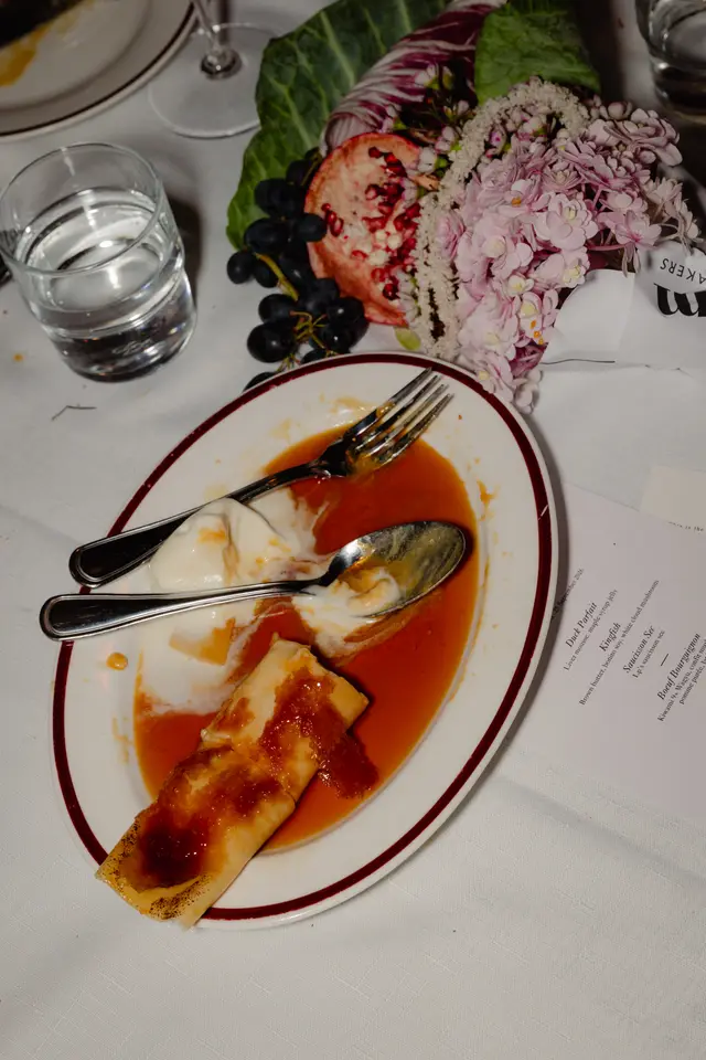 A top-down view of a partially eaten dessert on a white plate with a dark red rim, accompanied by a fork and spoon, resting on a white tablecloth. Beside it, a glass of water and a decorative centerpiece featuring pink flowers, grapes, pomegranate, and green leaves are visible, along with a printed menu.