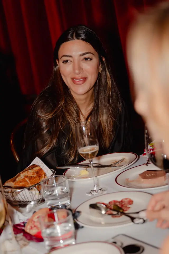 A candid portrait of a woman with long brown hair, smiling and looking towards the right, seated at a dining table. The table is set with plates, cutlery, glasses of beverages, bread, and other food items, suggesting a social event or dinner. A deep red curtain serves as a backdrop.