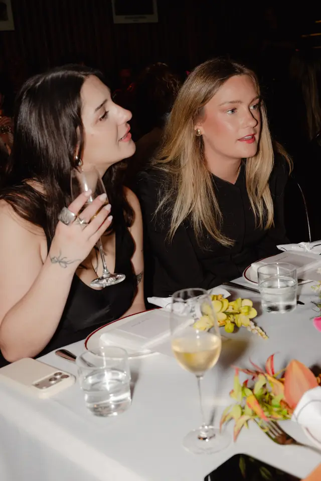 Two women are seated at a table during an evening event, engaged in conversation. The foreground shows a table setting with drinks, floral arrangements, and personal items. The background is dimly lit and blurred, suggesting a larger gathering.
