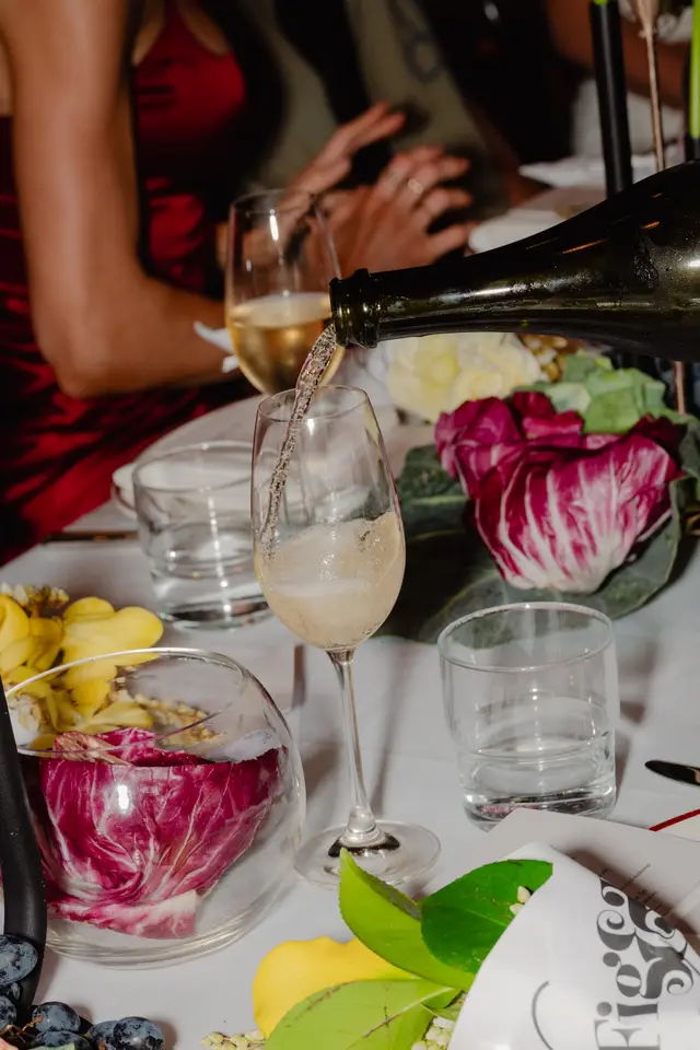A close-up shot captures sparkling wine being poured from a dark bottle into a tall glass, creating a stream of bubbles. The scene is set on a white tablecloth, adorned with a vibrant floral arrangement featuring purple radicchio, yellow petals, and green foliage. In the background, a person in a red dress and other hands are visible, suggesting a lively event.
