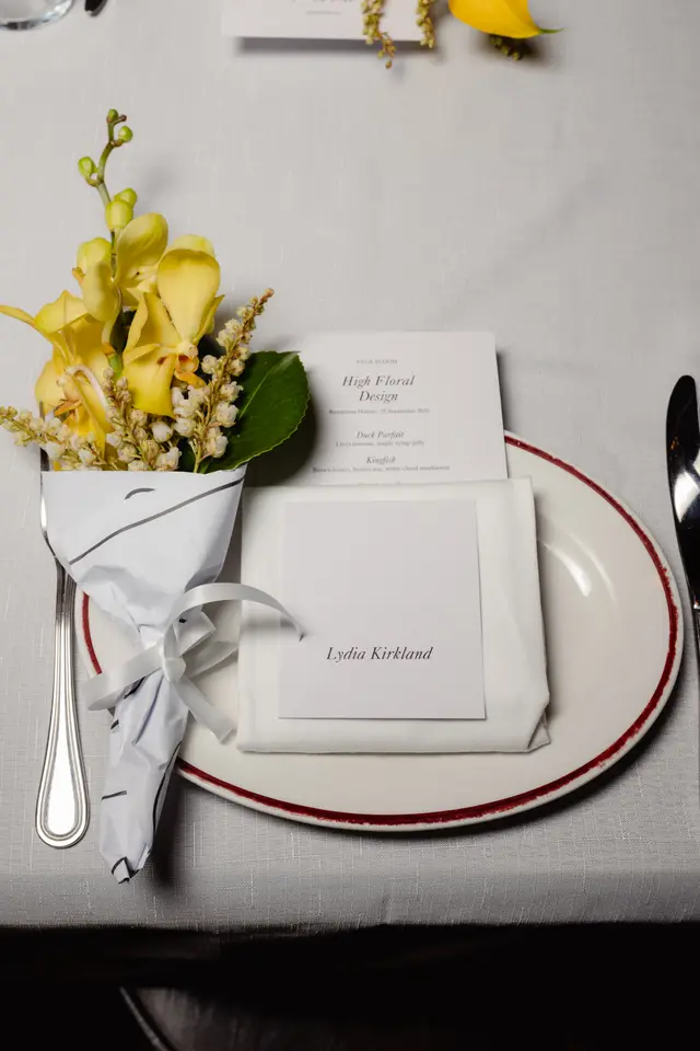 A close-up, overhead shot of an elegant place setting at an event. It features a small bouquet of yellow orchids and white flowers wrapped in paper, a menu card for 'High Floral Design' by Fig & Bloom, and a name card for 'Lydia Kirkland' on a white napkin, all resting on a white plate with a red rim on a light grey tablecloth.