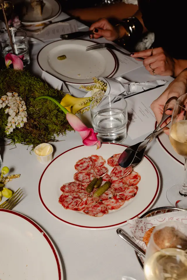 A high-angle, close-up shot of an elegantly set dining table at an event, featuring a white plate with sliced salami and gherkins being served with tongs. The table is adorned with a white tablecloth, fine dinnerware, and various floral arrangements including pink and yellow calla lilies, white bell-shaped flowers, and moss. Hands of multiple diners are visible, interacting with the food, cutlery, and a menu, suggesting an active and sophisticated dining experience.